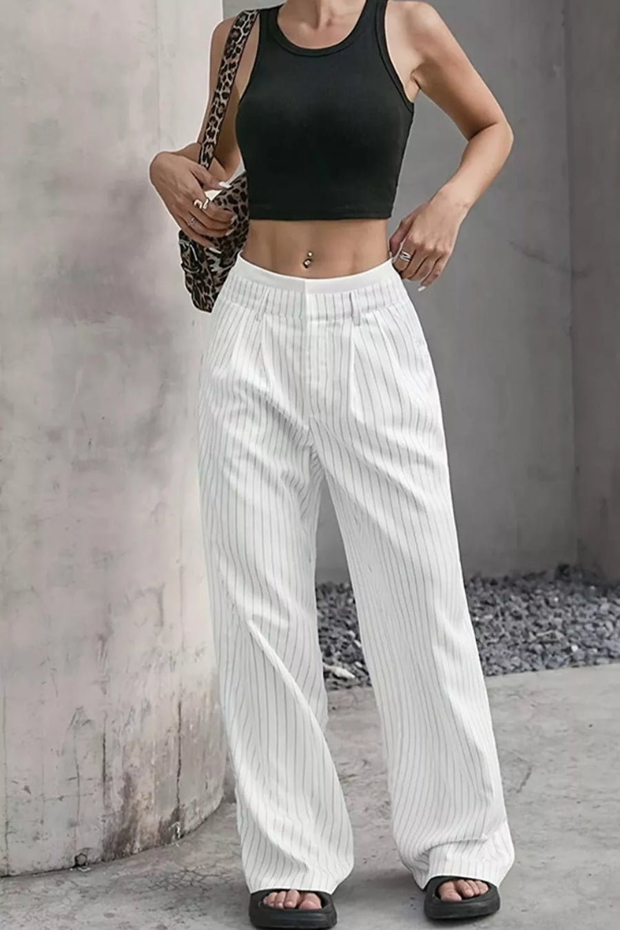 A woman wearing white wide-leg pinstripe pants with a black crop top, holding a leopard-print shoulder bag, standing against a textured concrete wall.