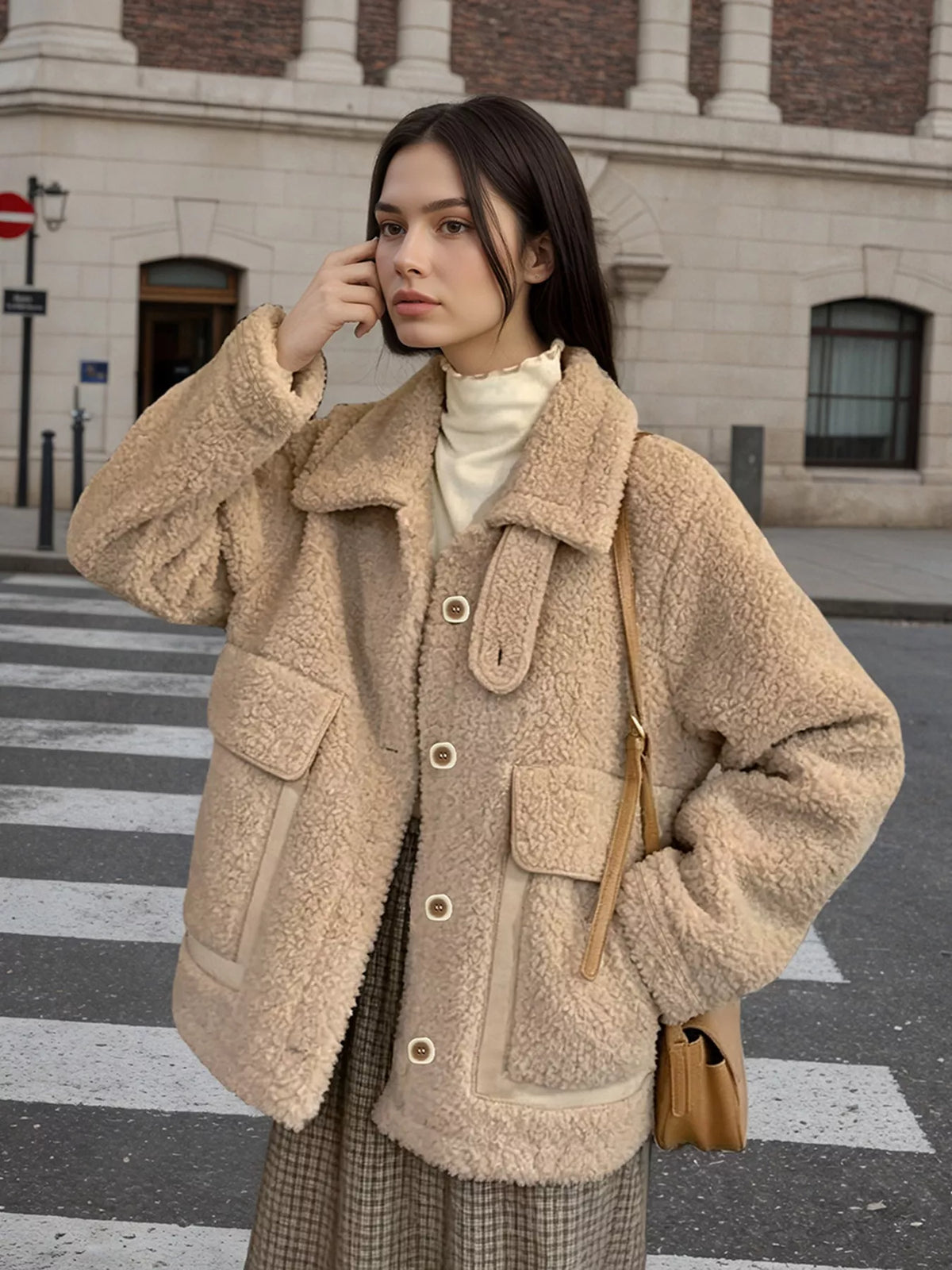 A woman stands on a city crosswalk wearing a beige sherpa fleece jacket over a cream turtleneck, with a brown plaid skirt and a tan shoulder bag. The background shows a blurred street scene with a stone building.