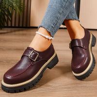 Brown leather shoes with buckles worn by a person on a wooden floor.