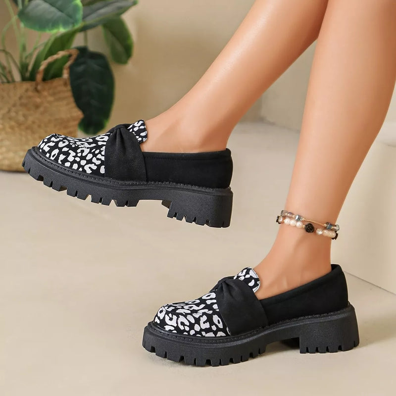 A person wearing black slip-on loafers with a black-and-white spotted fabric toe area, a black bow detail on the instep, and chunky lug soles. The photo is cropped to the lower legs and feet against a light beige indoor background with a plant visible in the upper left.