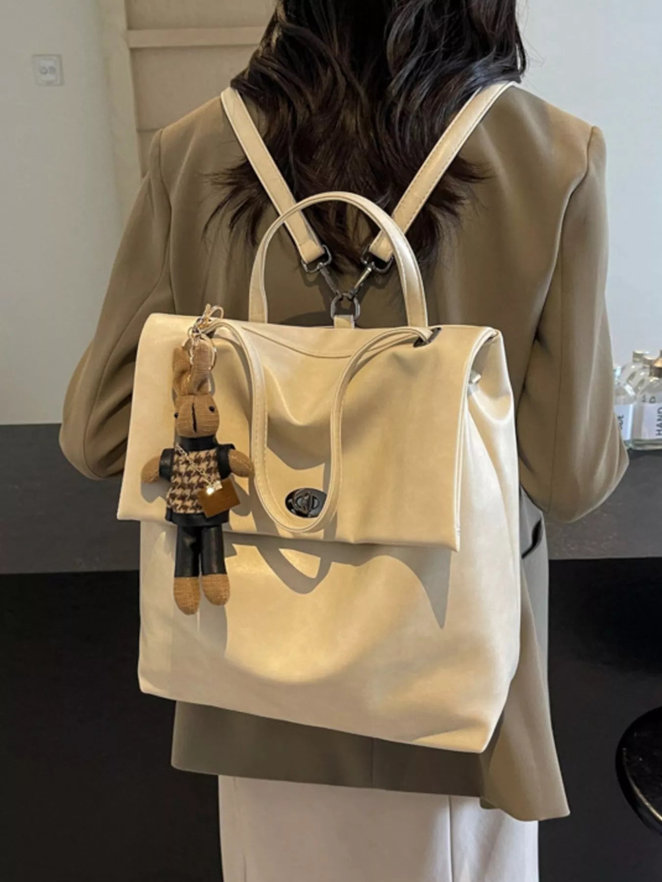 Person wearing a beige backpack with a small plush toy attached, standing indoors.