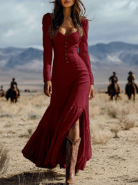Woman in a long burgundy dress and cowboy boots standing in a desert with people on horseback in the background.