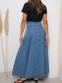 Person wearing a long blue denim skirt and black top against a white wall.
