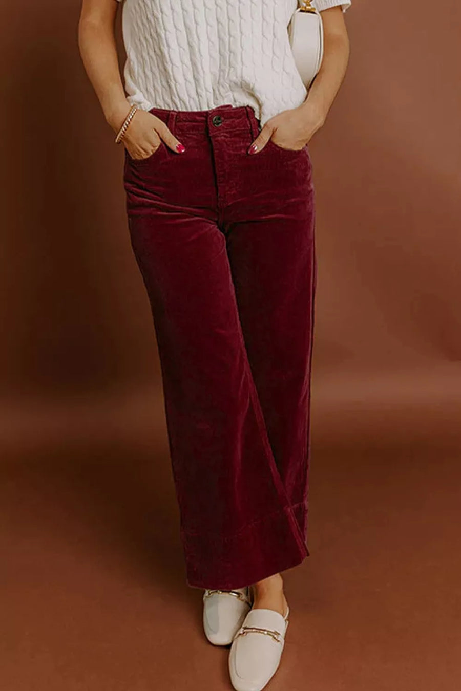 A person wearing burgundy wide-leg corduroy pants, a white knit top, and white loafers, standing against a warm brown studio backdrop.