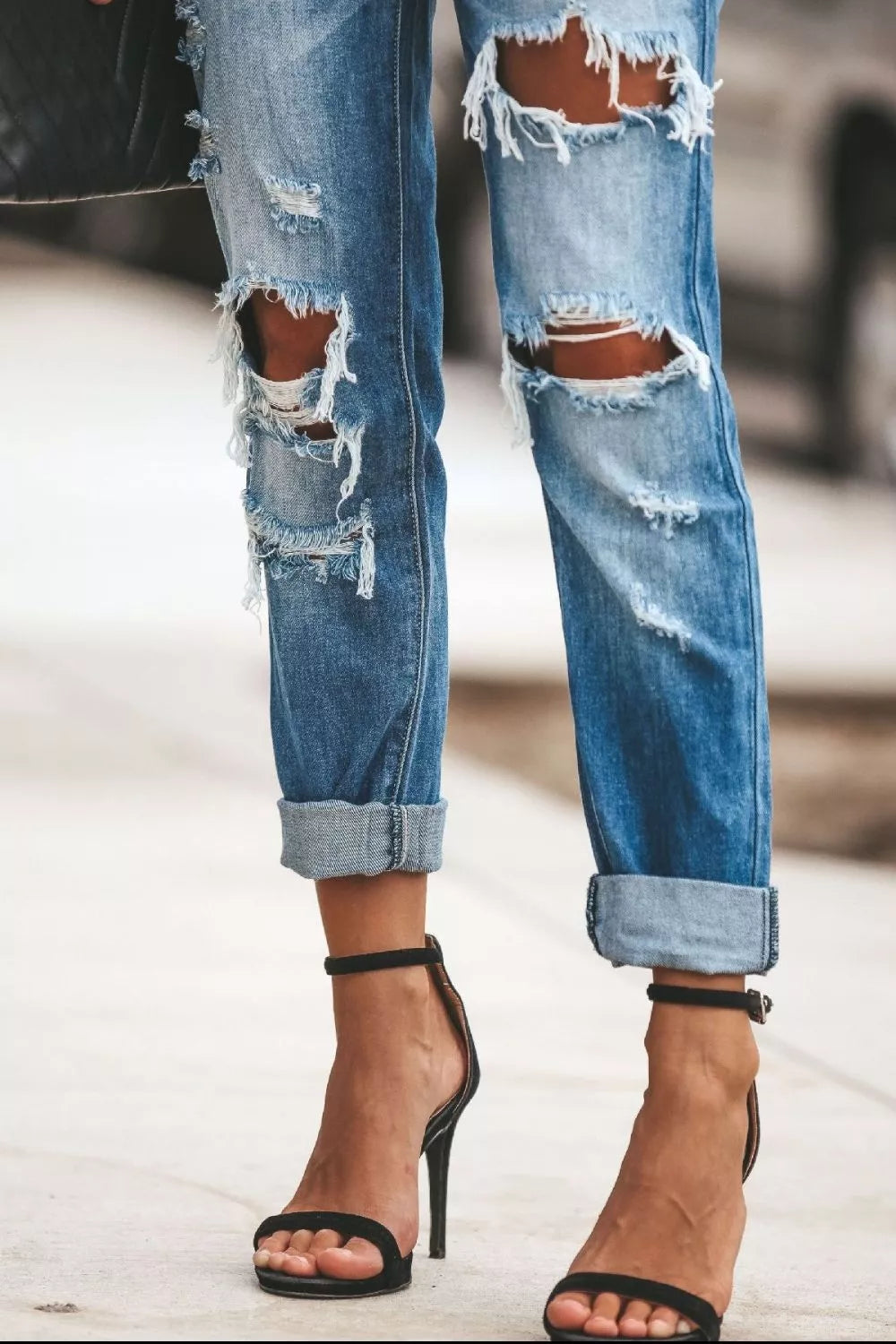 Ripped blue jeans with rolled-up cuffs worn with black high-heeled sandals on a blurred background.