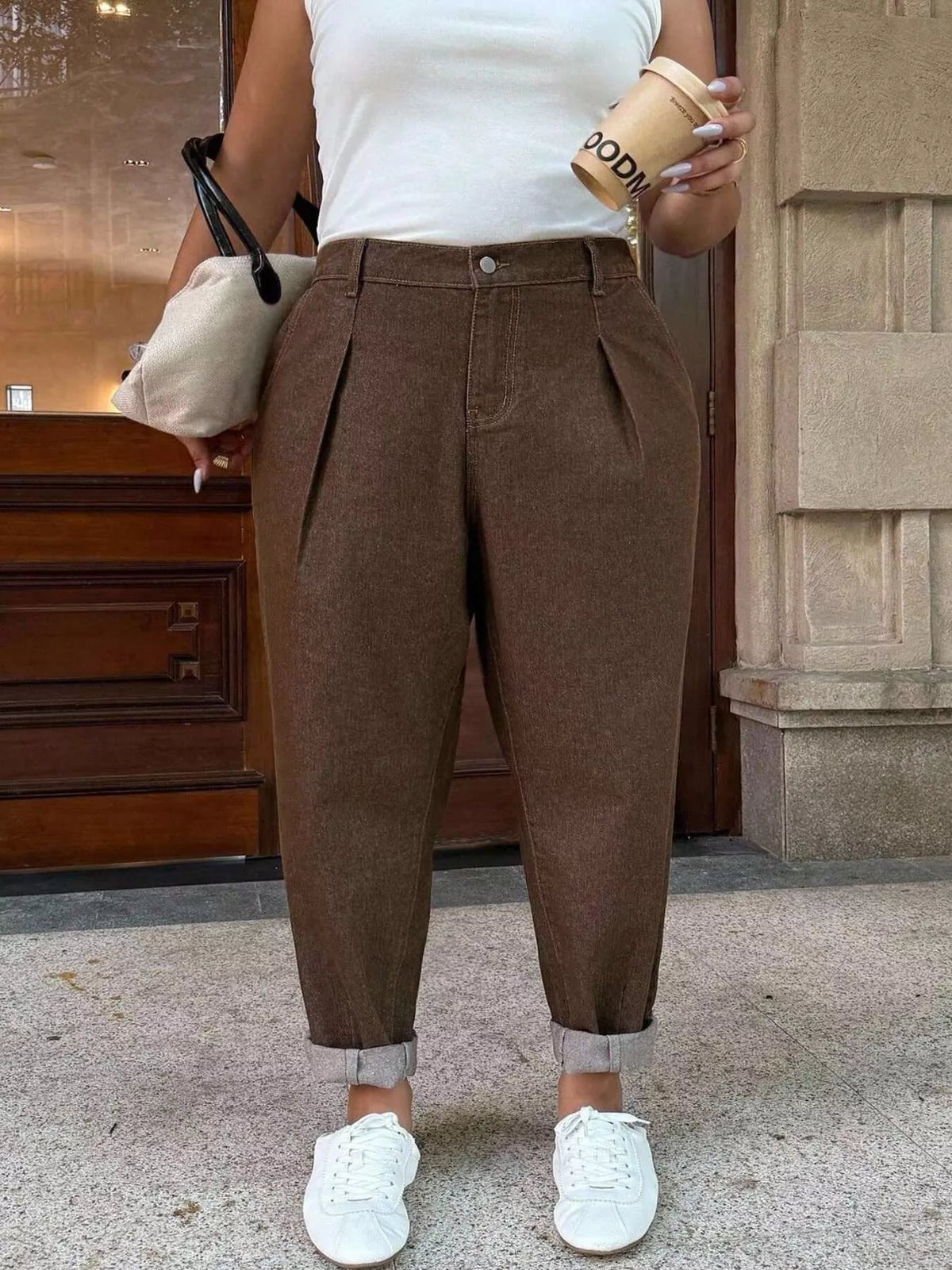 A person wearing brown pleated cuffed trousers, a white sleeveless top, and white sneakers, holding a beige handbag and a coffee cup in an outdoor urban setting.