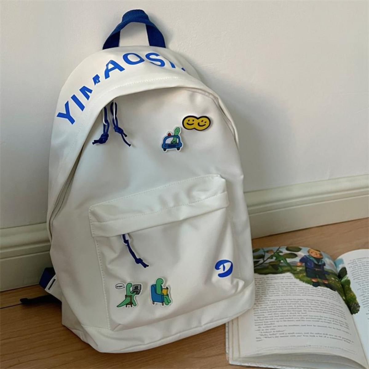 White backpack with colorful patches on a wooden floor next to an open book.