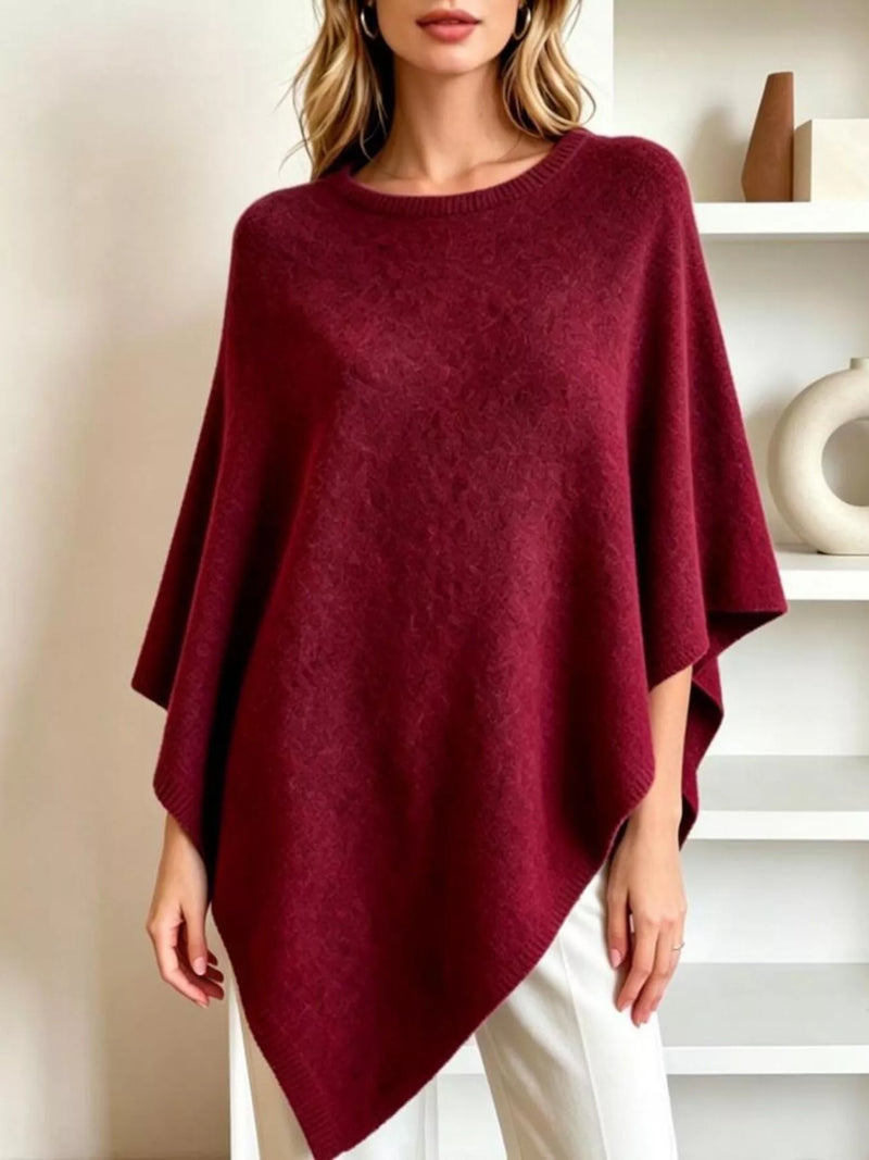 A woman wearing a deep burgundy knit poncho sweater with a crew neck and draped silhouette, paired with white pants, standing in a bright, modern room with white shelves.