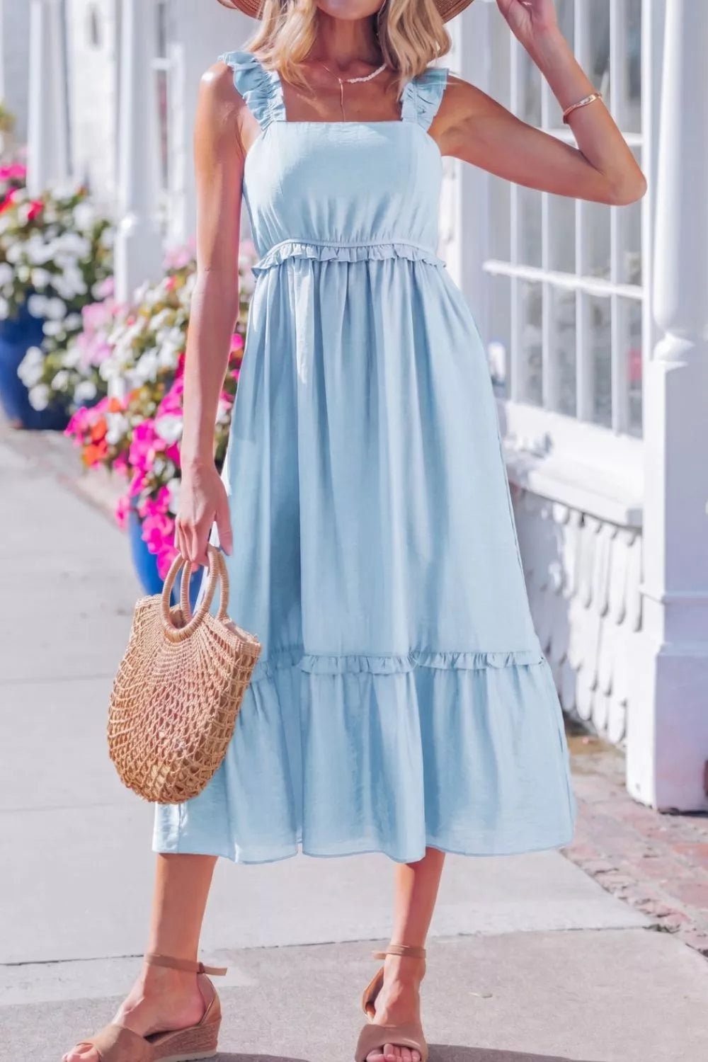 Woman wearing a light blue dress with ruffled sleeves and bottom, holding a straw bag on a sunny day.