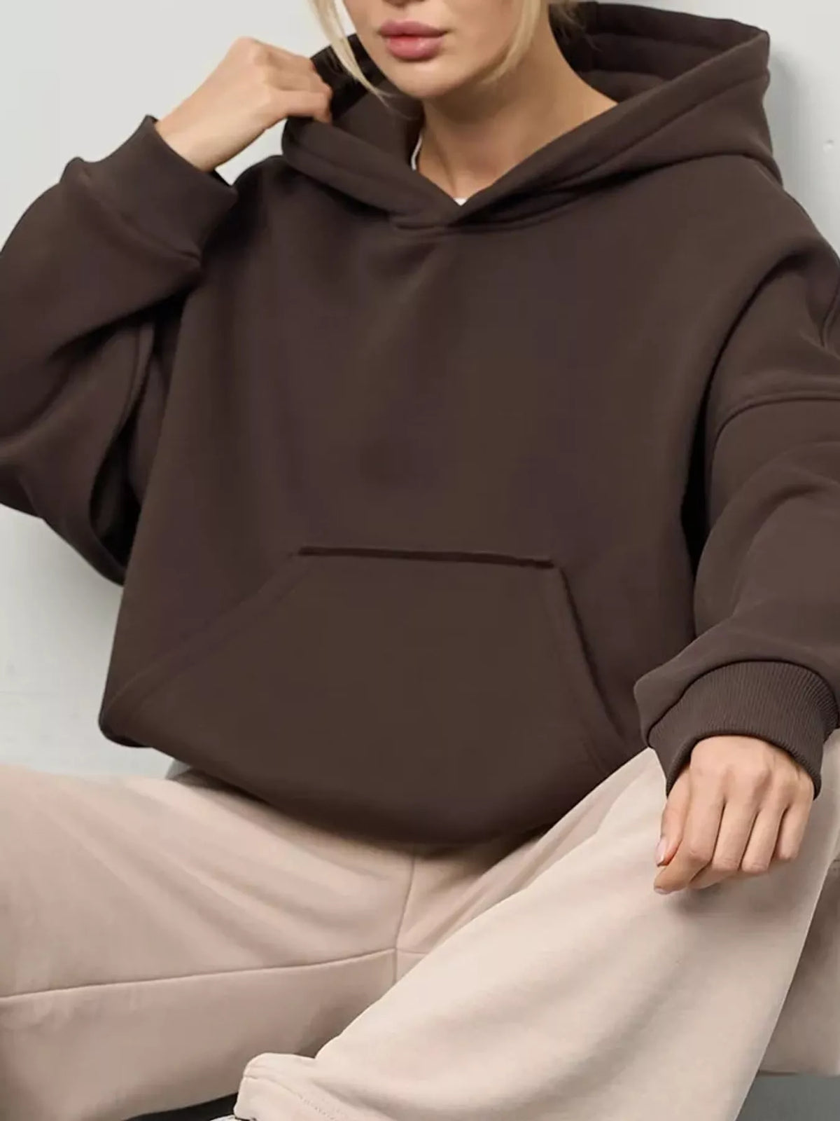 A woman wearing a dark brown oversized pullover hoodie with a front kangaroo pocket, paired with light beige sweatpants, shown in a close-up lifestyle shot.