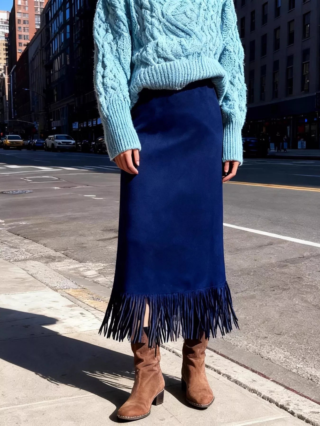 Person wearing a blue sweater, blue fringed skirt, and brown boots on a city street.