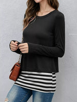 A woman wearing a black long-sleeve crewneck top layered over a black and white striped tunic, paired with blue jeans and carrying a small brown crossbody bag against a textured gray wall.