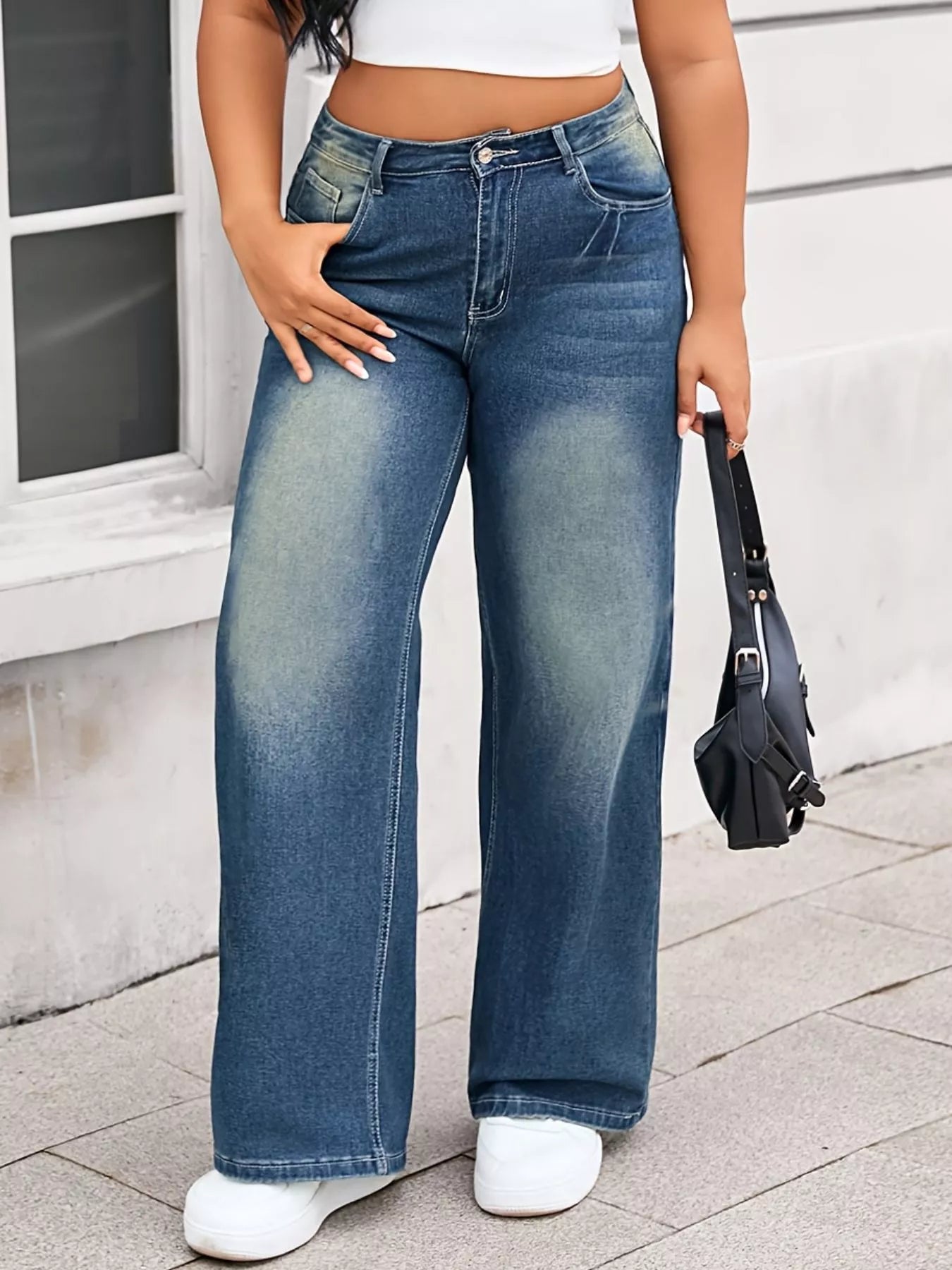 Person wearing blue jeans and white sneakers standing on a sidewalk.