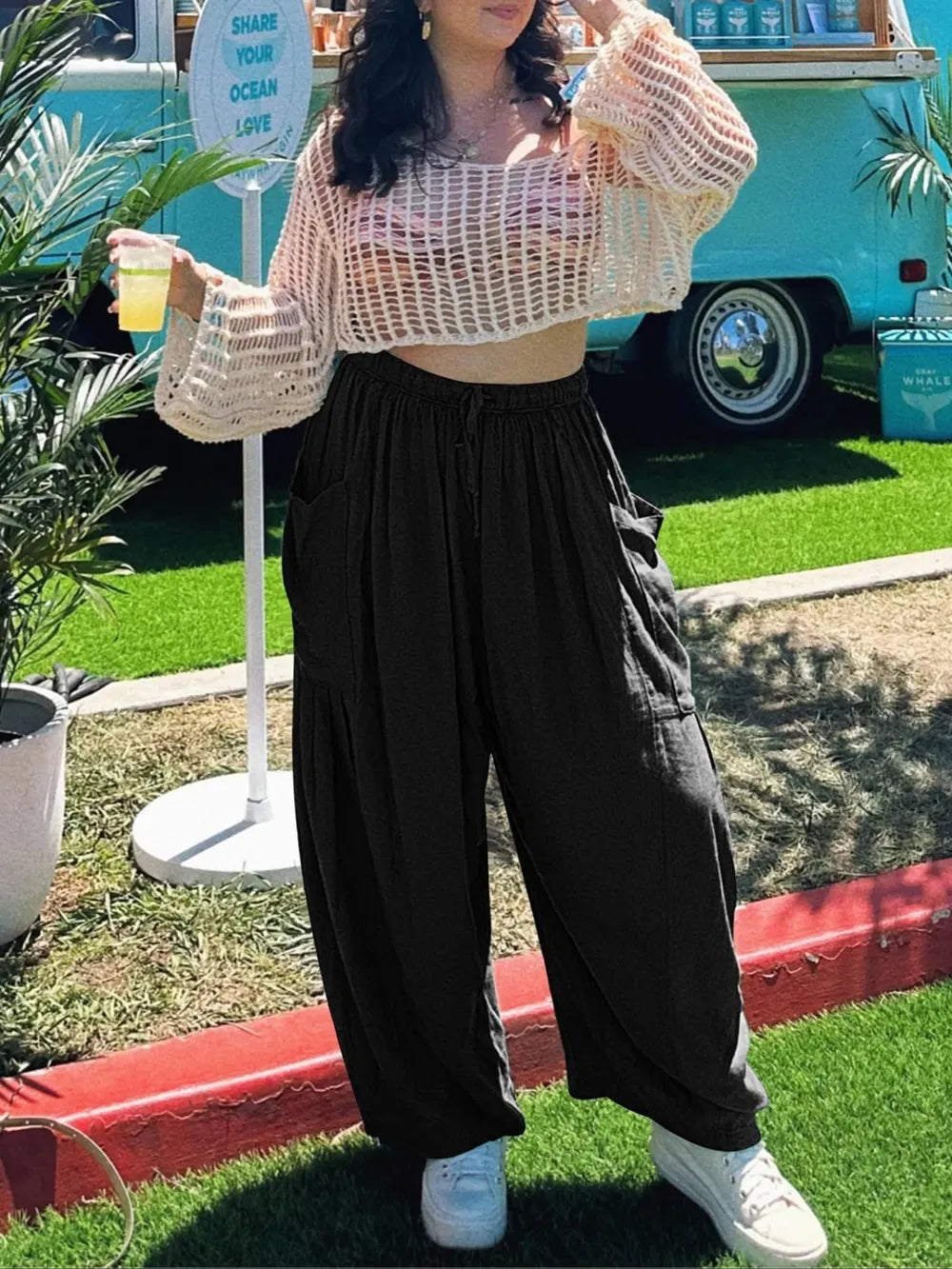 Person wearing a beige crochet top and black pants standing outdoors with a colorful vehicle in the background.