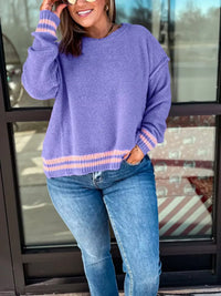 Person wearing a purple sweater with striped sleeves and blue jeans standing in front of a glass door.