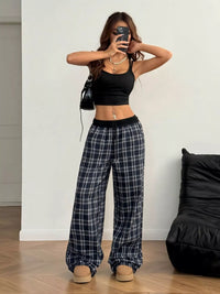 Woman taking a mirror selfie wearing a black crop top and plaid pants in a minimalistic room.
