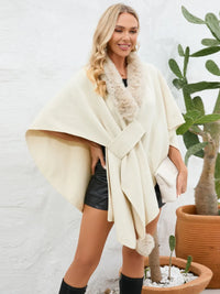 Woman wearing a cream-colored poncho with fur trim, standing against a white wall with plants.