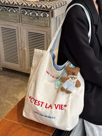 White tote bag with 'Cést La Vie' text and teddy bear charm, held by a person.