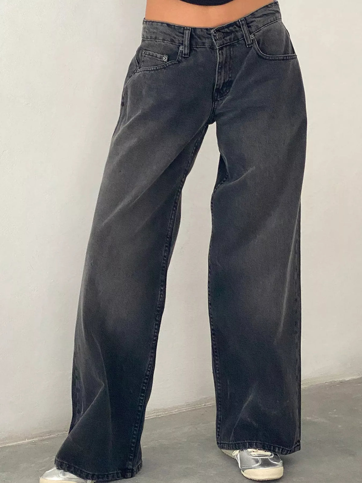 A person wearing dark gray wide-leg jeans with a faded wash, paired with white sneakers, standing against a plain light gray wall.
