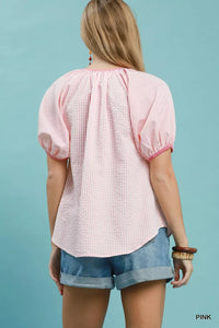 Woman wearing a pink checkered blouse and denim shorts against a light blue background