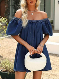 Woman wearing a blue off-shoulder dress holding a white handbag outdoors.