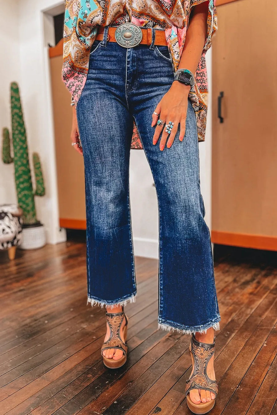 Person wearing blue jeans and a patterned top with a belt, standing on a wooden floor.
