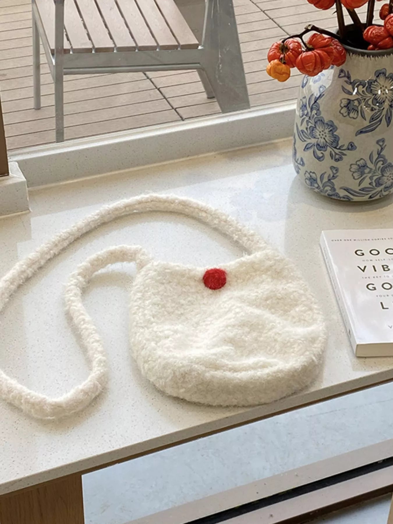White textured handbag with a red button on a light surface, with a vase and books in the background.
