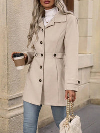 Woman in a beige coat holding a coffee cup and a handbag, standing against a stone wall.