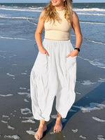 Woman standing on a beach wearing a beige top and white pants.