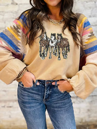 Person wearing a beige sweater with horse design and colorful sleeves, paired with blue jeans against a neutral background.