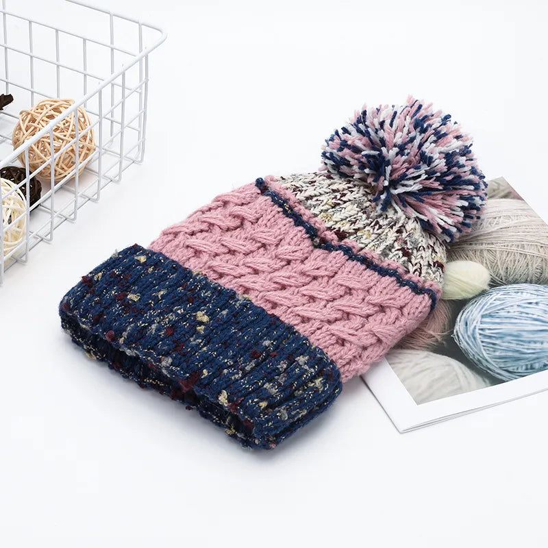 Knitted beanie with pom-pom on a white surface with yarn balls and a basket in the background.