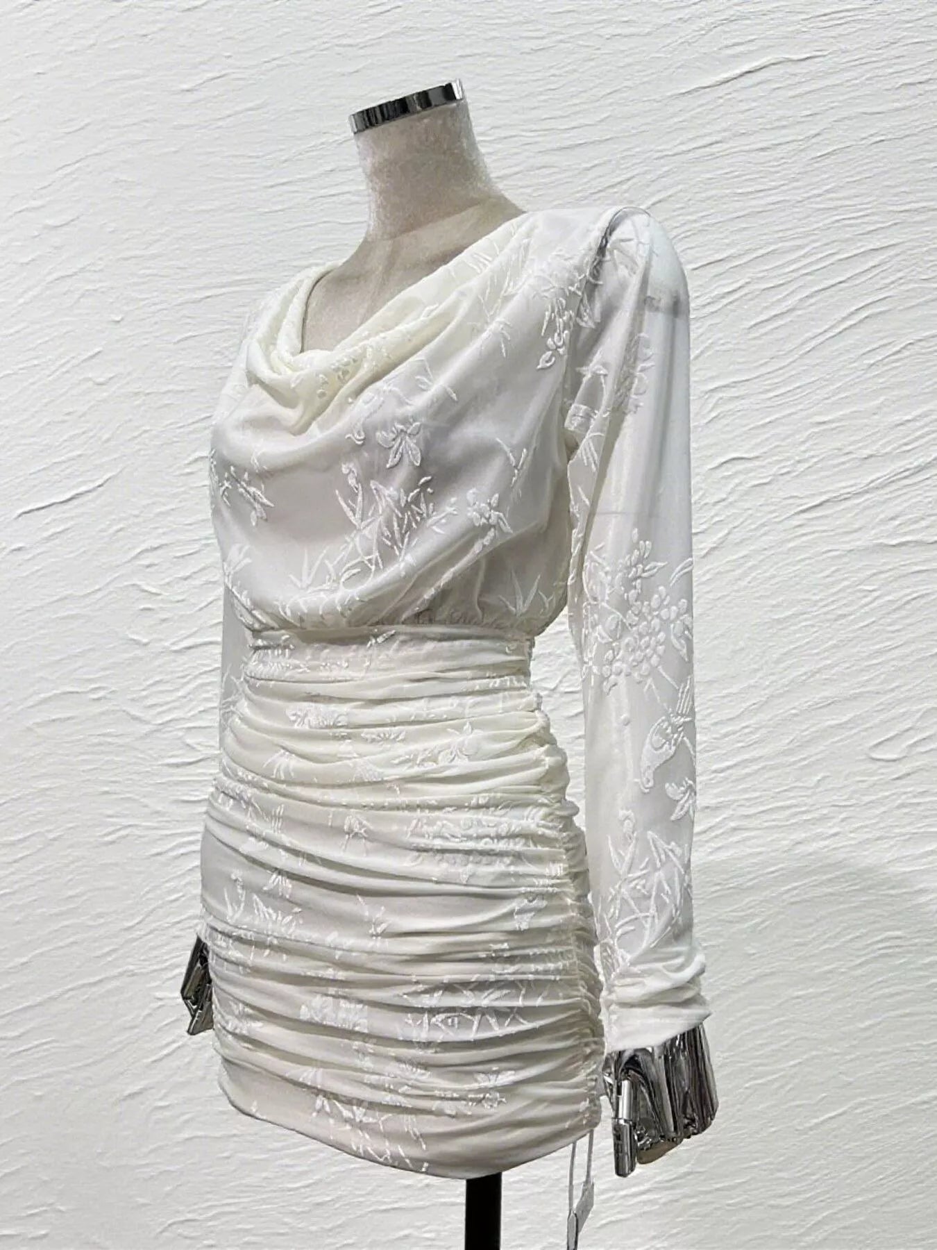 An off-white ruched mini dress with a draped cowl neckline and long sheer sleeves featuring white floral embroidery, displayed on a mannequin against a textured white wall.
