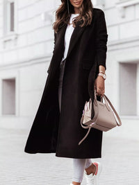 Person wearing a black coat and white pants, holding a beige handbag against a light-colored wall.