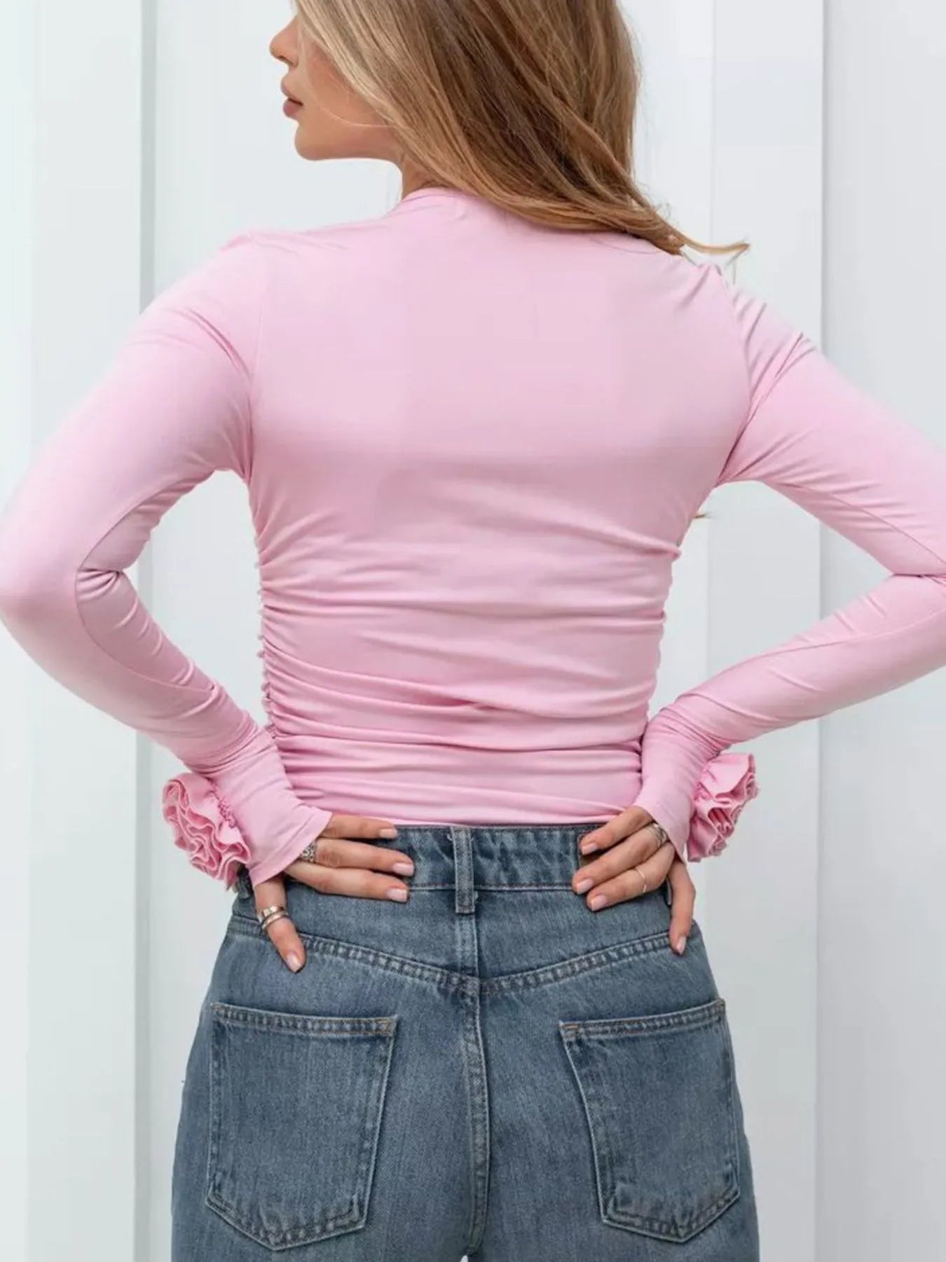 Person wearing a pink long-sleeve top with ruffled cuffs and blue jeans against a white background
