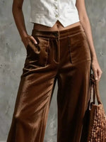 A person wearing high-waisted wide-leg brown velvet pants with a white cropped vest top, holding a woven brown bag against a textured gray wall.