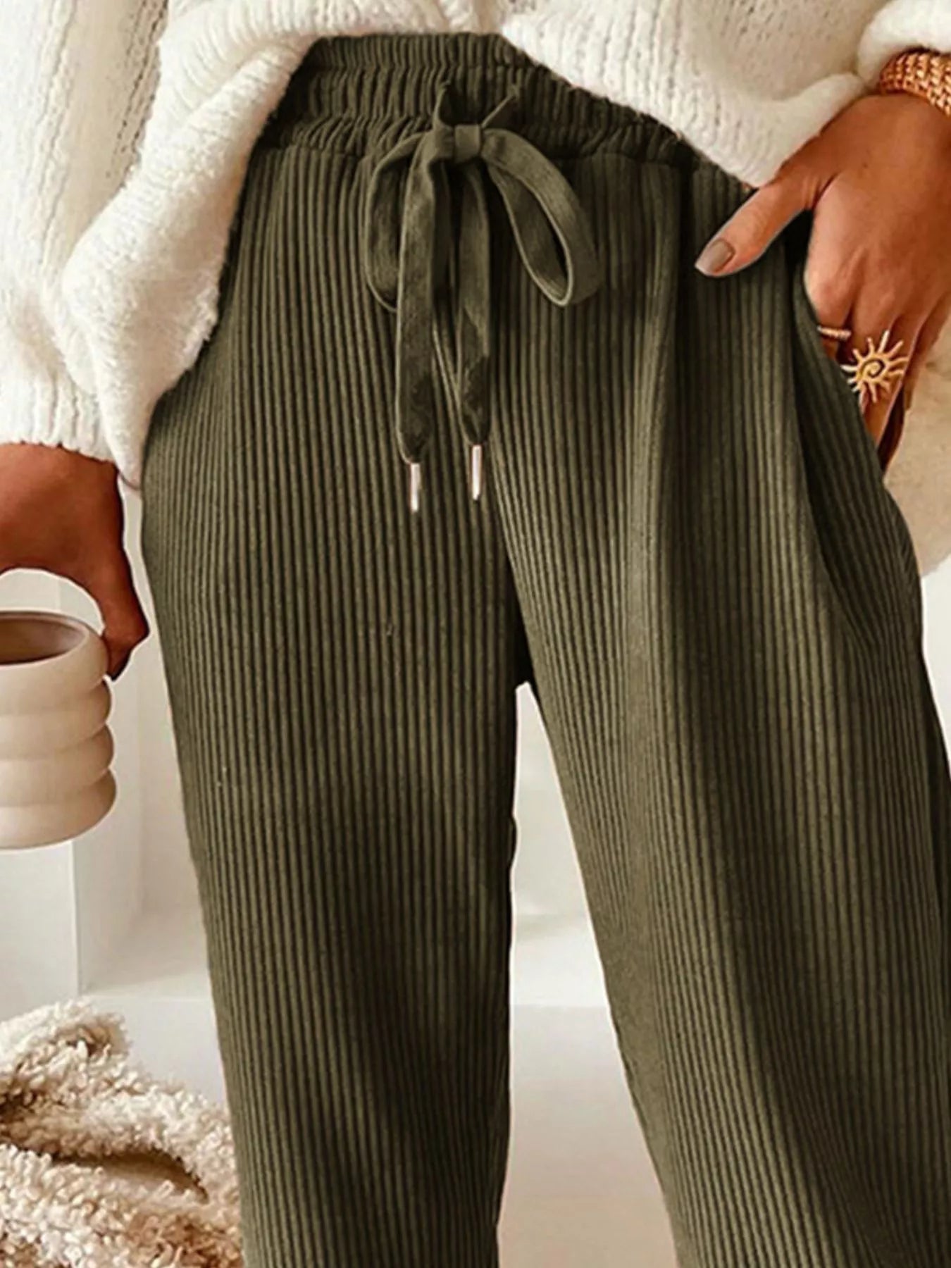 Person wearing olive green ribbed pants with a white sweater and beige background