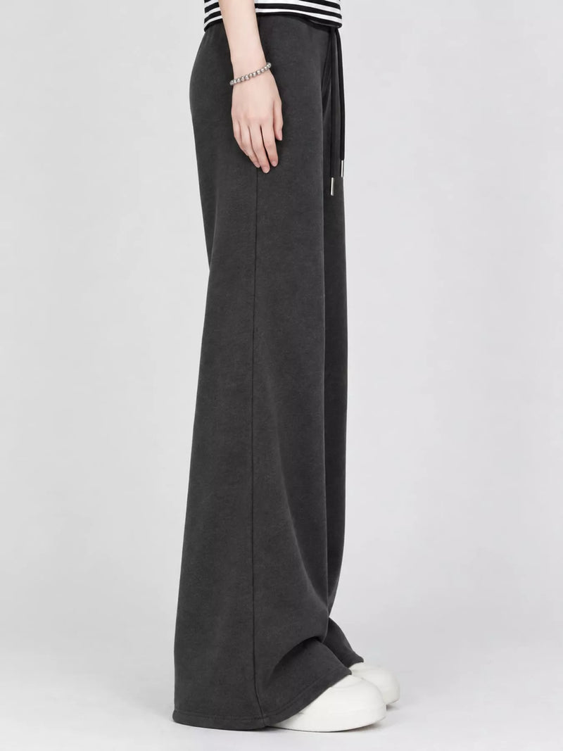 A person wearing dark charcoal gray wide-leg knit pants with a drawstring waist, paired with white slip-on shoes, shown from the waist down against a light gray studio background.