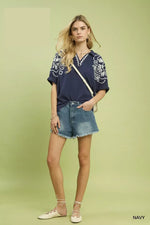 Woman wearing a navy blue top with white patterns and denim shorts on a light green background