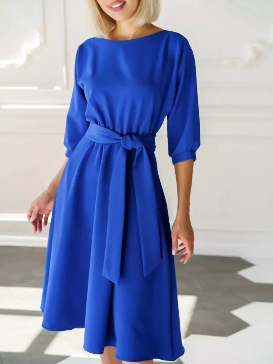 A woman wearing a bright royal blue midi dress with three-quarter sleeves, a boat neckline, and a matching fabric tie belt at the waist, standing in a bright white interior.