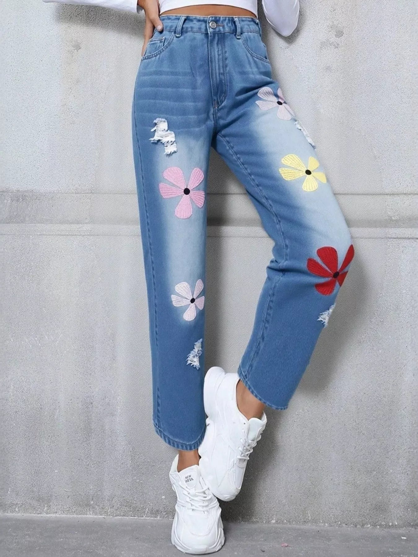 Blue jeans with colorful flower designs worn by a person against a gray wall.