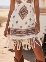 Person wearing a white dress with tribal patterns and fringes, standing on a beach.