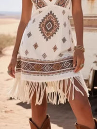 Person wearing a white dress with tribal patterns and fringes, standing on a beach.