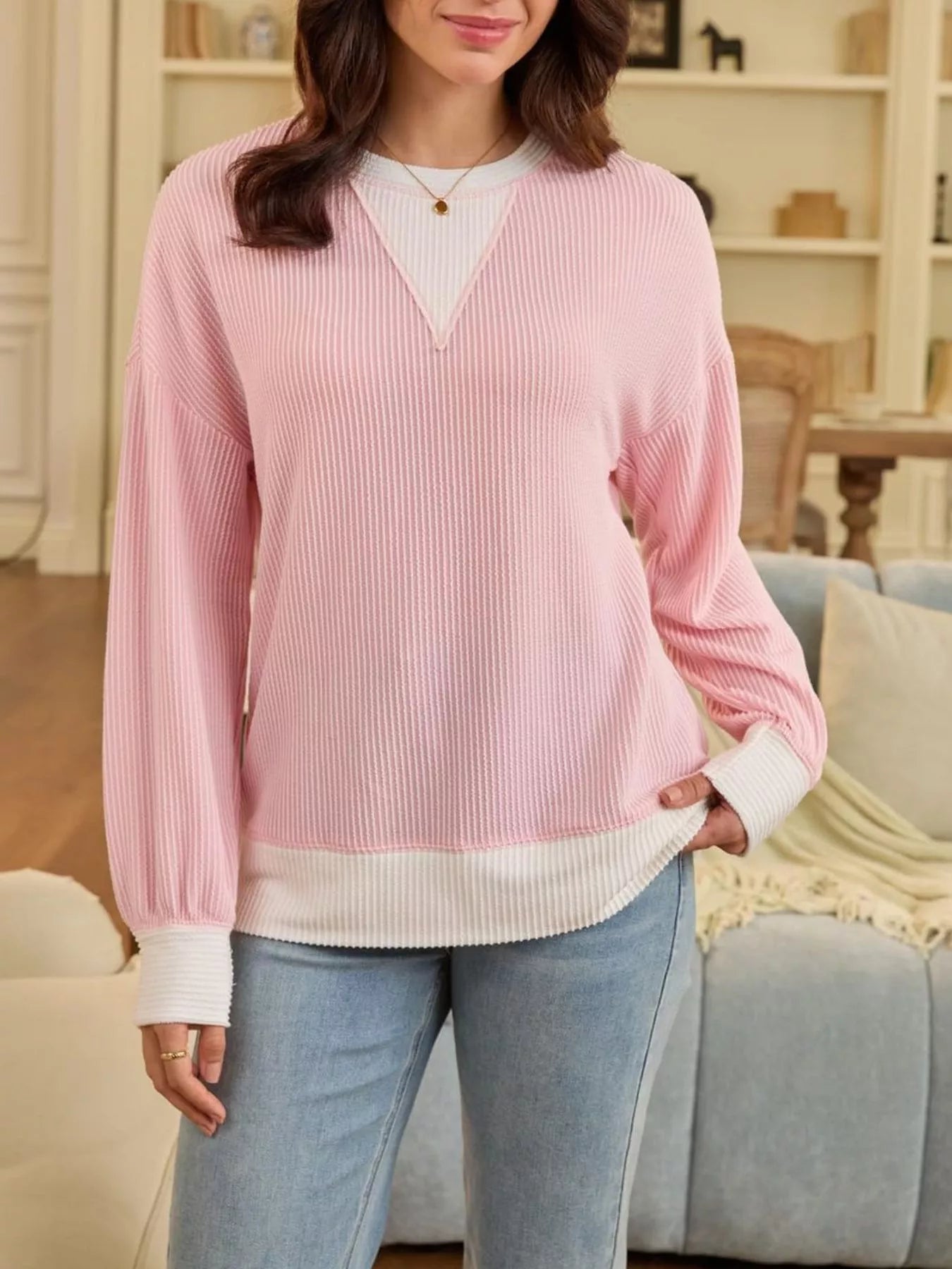 Woman wearing a pink ribbed sweater with a white shirt underneath, standing in a living room.
