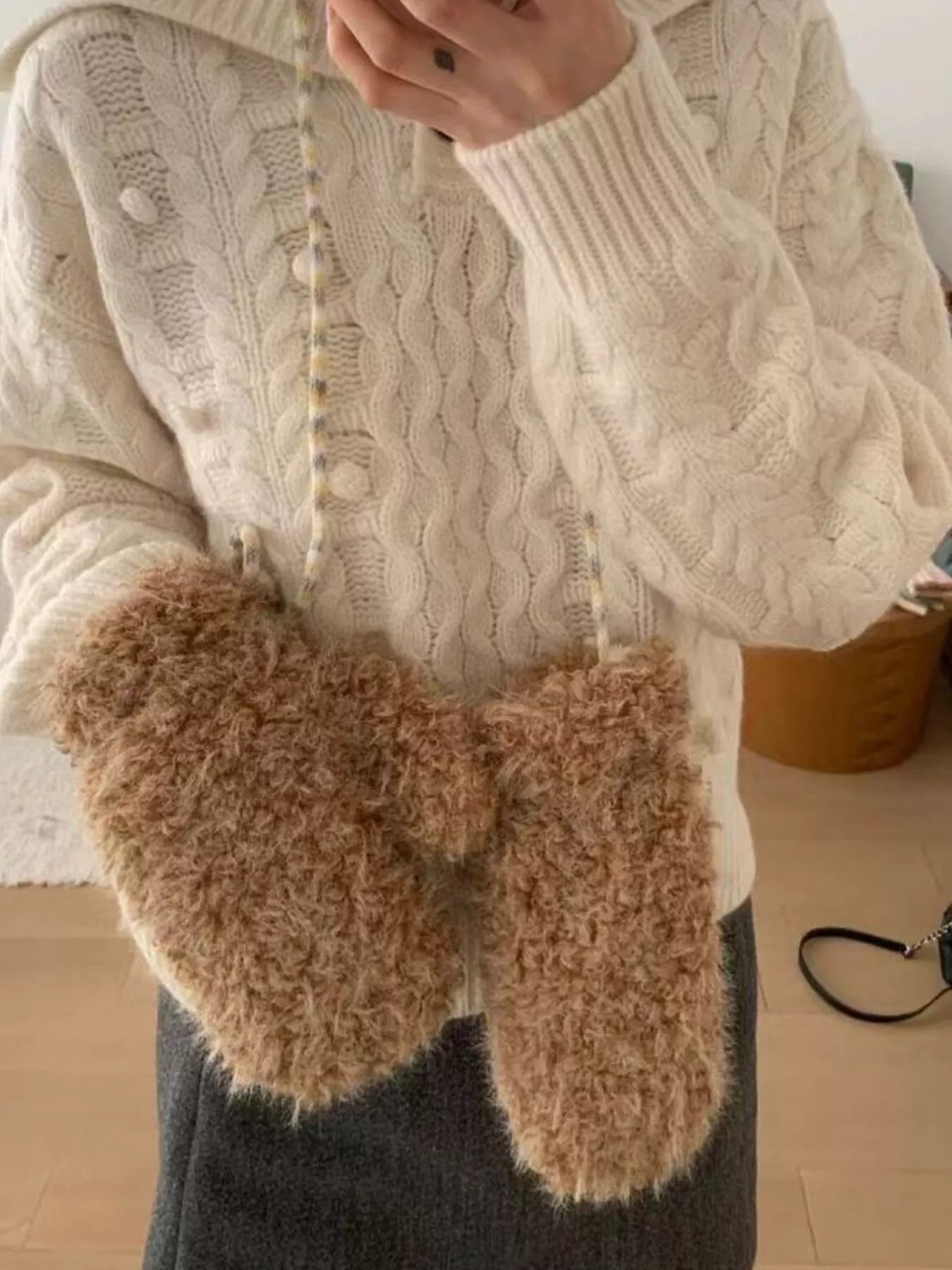 Person wearing a white cable knit sweater and brown fuzzy gloves indoors.