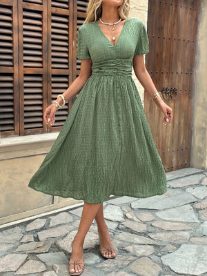 Woman wearing a green dress standing on a stone pavement with wooden shutters in the background.