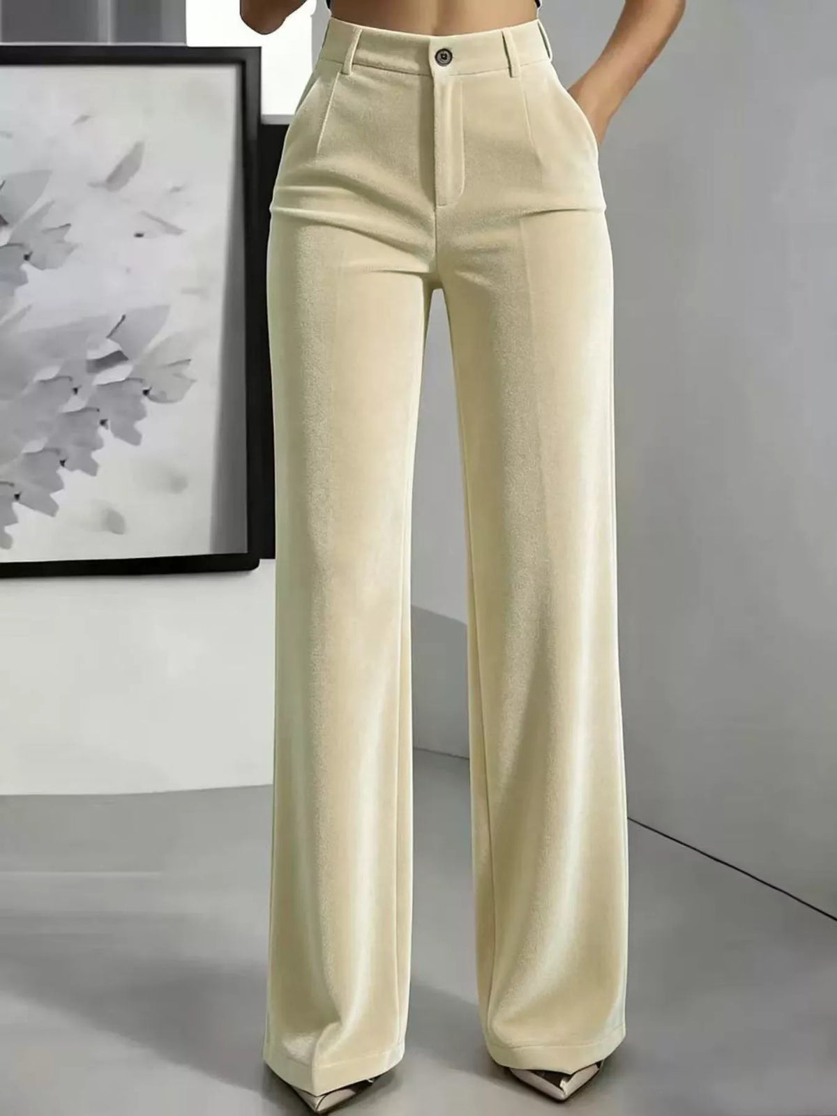 A model is wearing a pair of high-waisted, wide-leg trousers in a light cream or off-white color. The pants feature a structured waistband with belt loops, a button closure, and a straight, flowing leg.