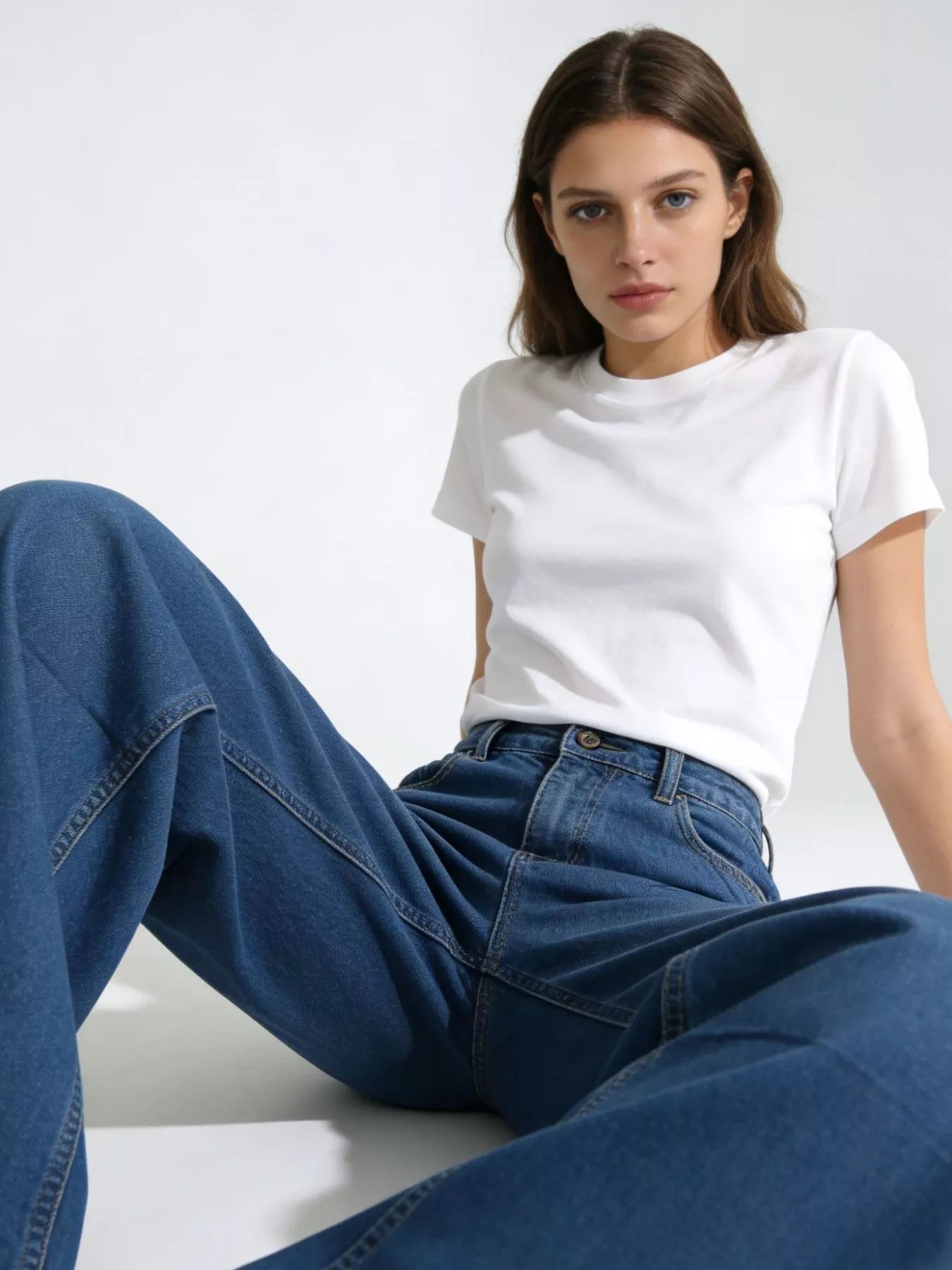 Woman wearing a white t-shirt and blue jeans sitting on a white surface with a white background