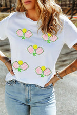 Person wearing a white t-shirt with pickleball paddle designs outdoors.