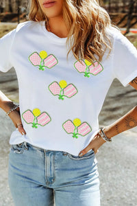 Person wearing a white t-shirt with pickleball paddle designs outdoors.