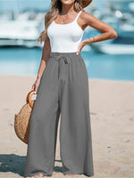 Woman in white tank top and gray wide-leg pants standing on a beach with boats in the background.